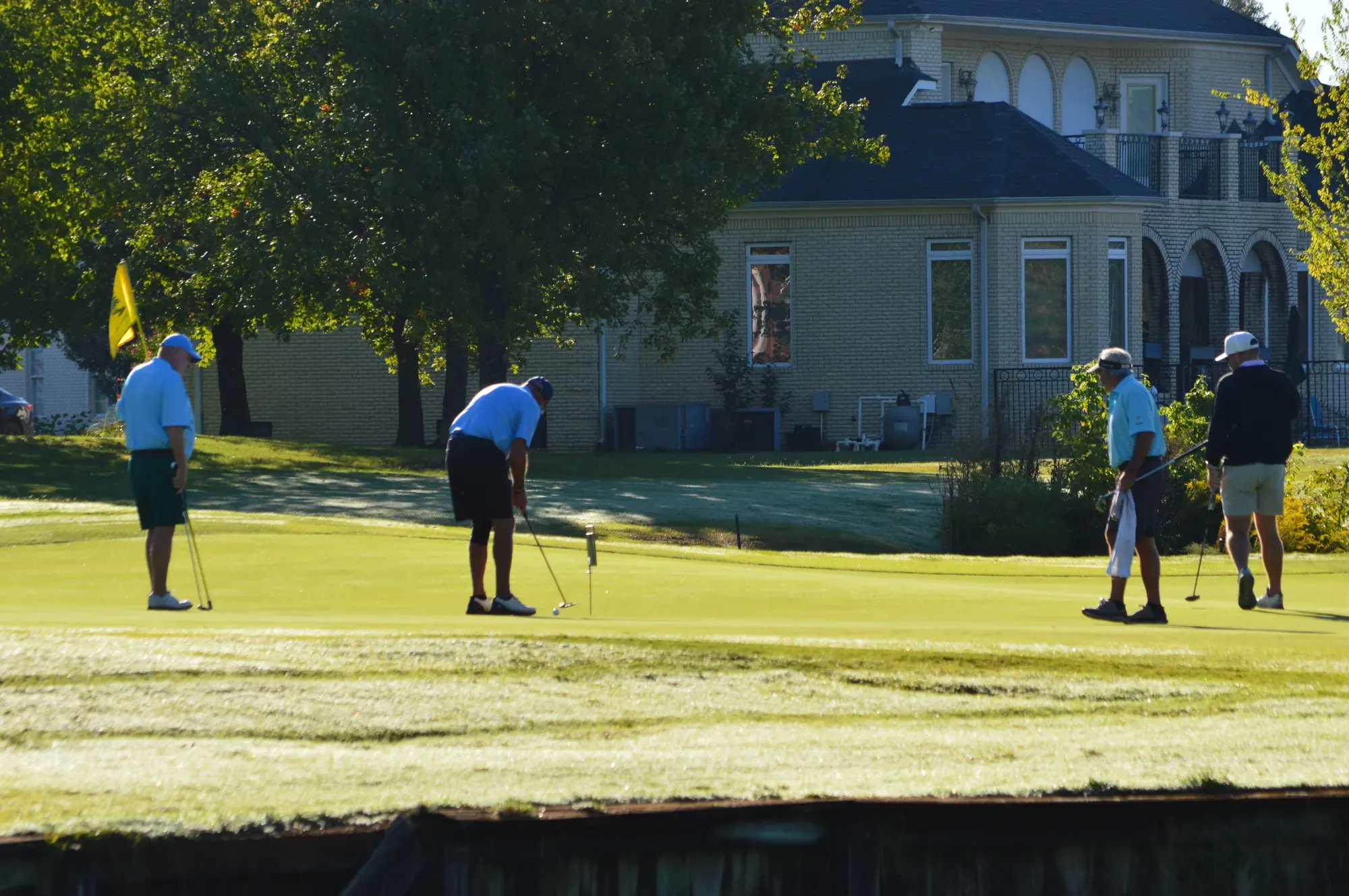 Four golfers on a putting green with a yellow flag, with a brick clubhouse behind them