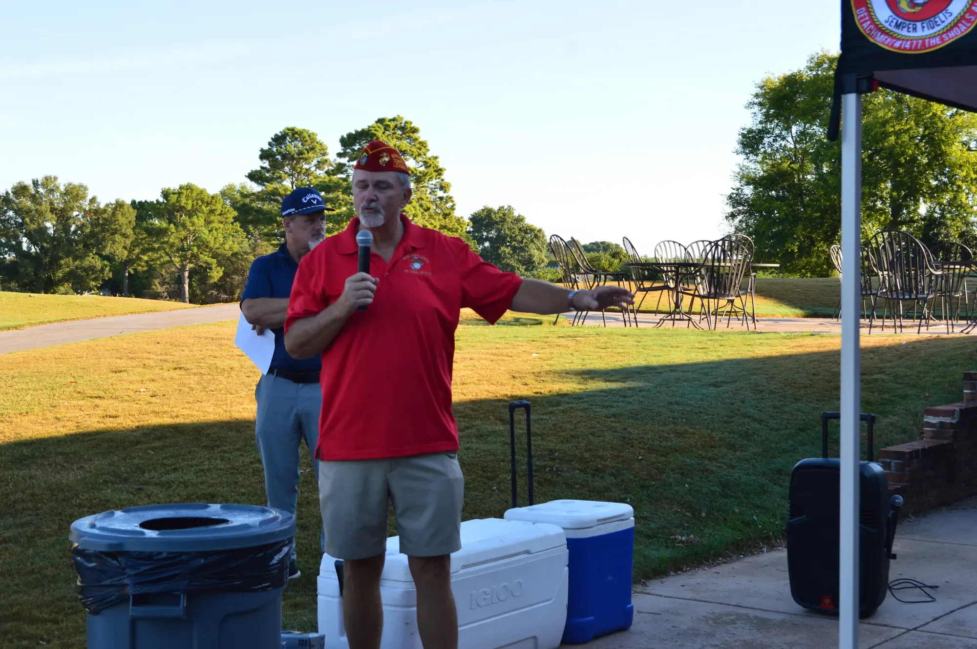 A detachment member in a red Marine Corps League shirt speaks into a microphone to the assembled players